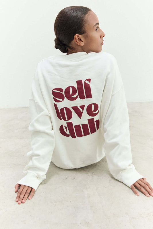 Person wearing a white sweatshirt with 'self love club' text on the back, sitting on a light-colored floor.