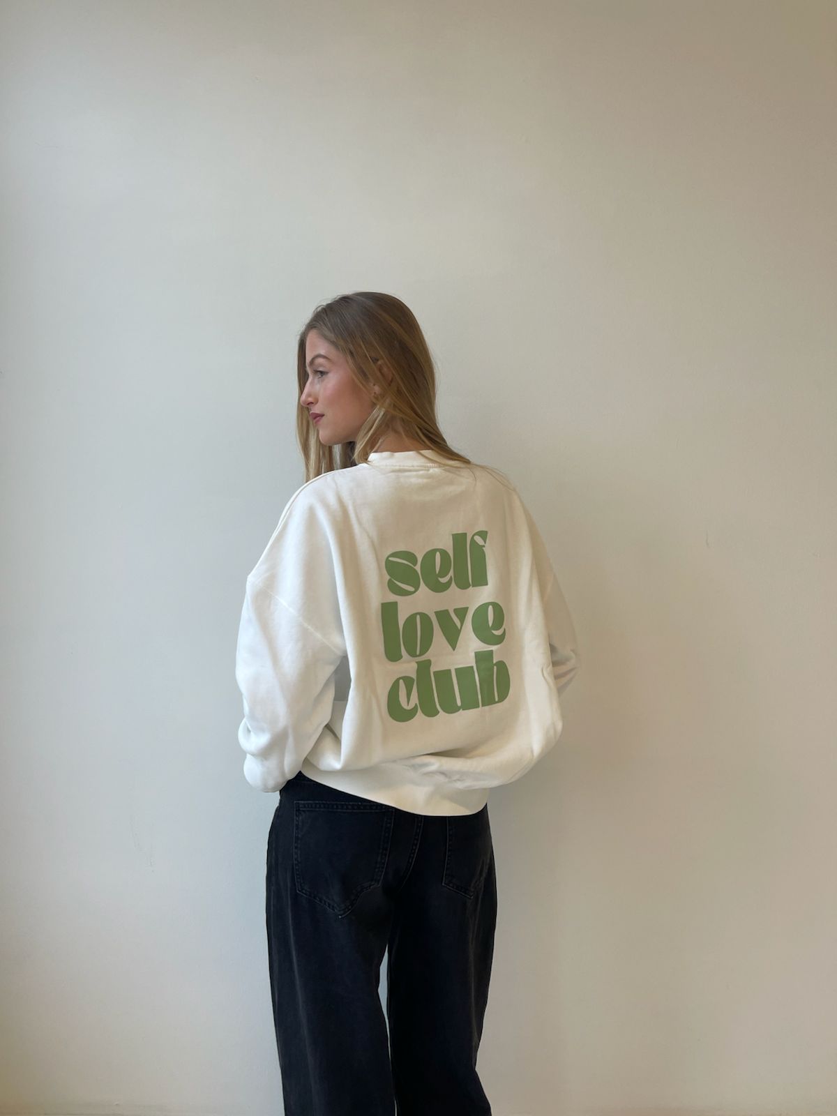 Person wearing a white sweatshirt with 'self love club' text on the back against a plain wall.