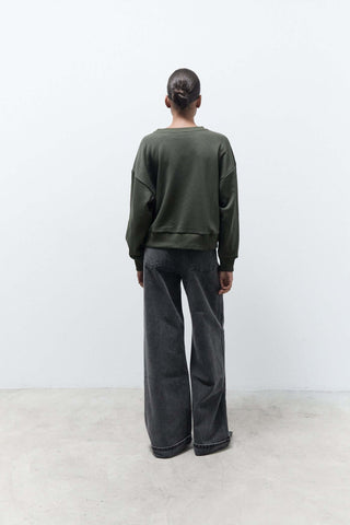 Person wearing a green sweater and dark jeans standing against a white wall.