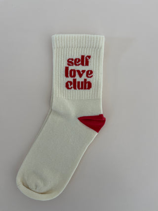 A pair of white socks with the text 'self love club' printed in red on the cuff, along with red accents on the heel and toe.
