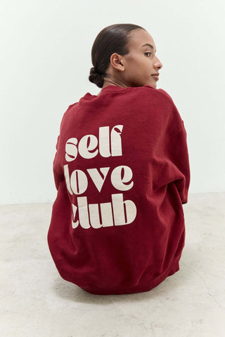 Person wearing a red sweatshirt with 'self love club' text on the back against a white background