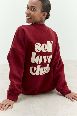 Person wearing a maroon sweatshirt with 'self love club' text on a light background