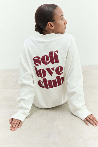 Person wearing a white sweatshirt with 'self love club' text on the back, sitting on a light-colored floor.