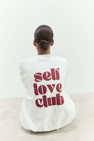 Person wearing a white sweatshirt with 'self love club' text on the back, sitting on a light-colored floor.
