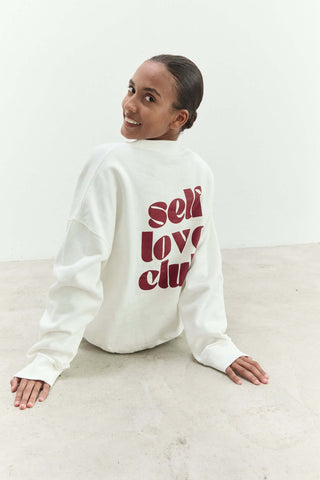Person wearing a white sweatshirt with 'self love club' text on a light background
