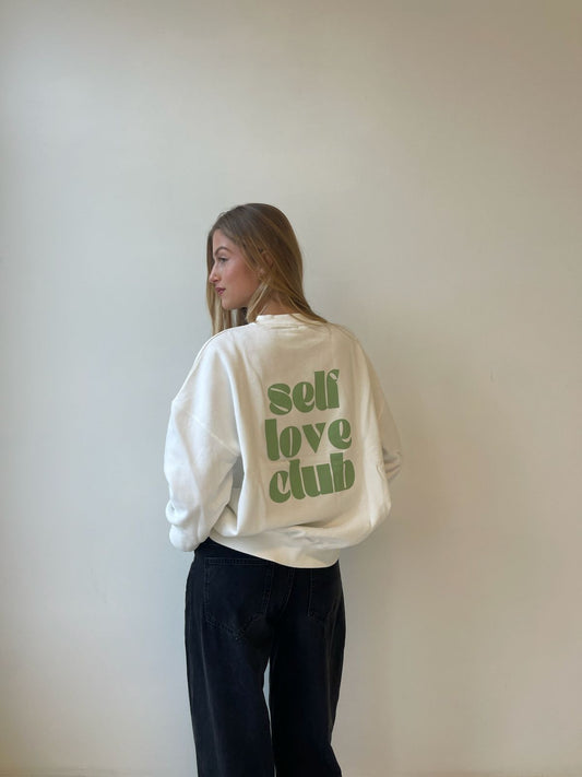 Person wearing a white sweatshirt with 'self love club' text on the back against a plain wall.